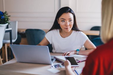 Çok ırklı gülümseyen, dizüstü bilgisayarla çalışan ve akıllı telefonla sörf yapan genç bayanlar modern ışık yaratıcı çalışma alanında masada oturuyorlar.