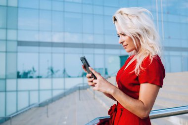 Aydınlık bir günde bulutları yansıtan modern cam binadaki merdivenlerde parlak kırmızı elbiseli düşünceli bir kadının cep telefonu sörfü görüntüsü.