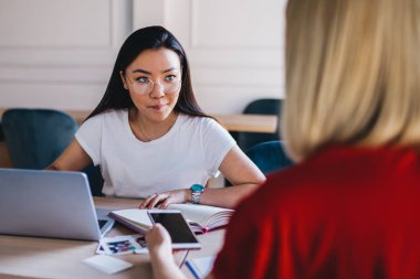 Çok ırklı gülümseyen, dizüstü bilgisayarla çalışan ve akıllı telefonla sörf yapan genç bayanlar modern ışık yaratıcı çalışma alanında masada oturuyorlar.
