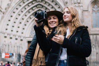 Sıcak giysili neşeli yetişkin kadınlar ellerinde fotoğraf makinesiyle eski katedrale karşı durup gözlerini kaçırırken fotoğraf çekiyorlar.