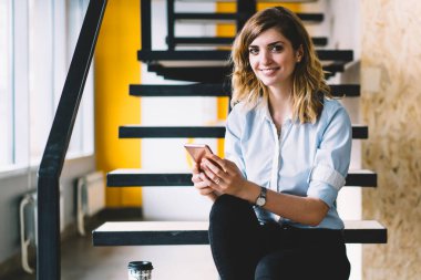 Ofis merdivenlerinde cep telefonu ile otururken kameraya bakan mutlu gülümseyen kadın çalışanın portresi, online alışveriş ve alışveriş için hücresel uygulama kullanarak resmi giyim pozitif kadın