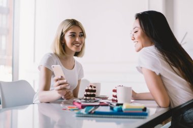 Kafe masasında birlikte oturan, cep telefonu görünümlü ve gülen iki kadının yan görüntüsü.