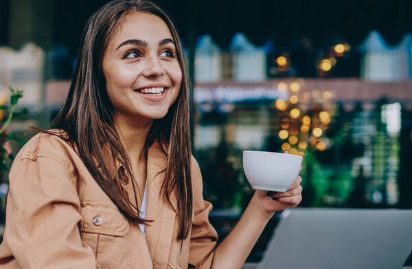 Cheerful brunette female hipster enjoying aroma coffee break holding cup and looking away smiling, skilled successful woman freelancer doing remote job on laptop computer spending time at cafe