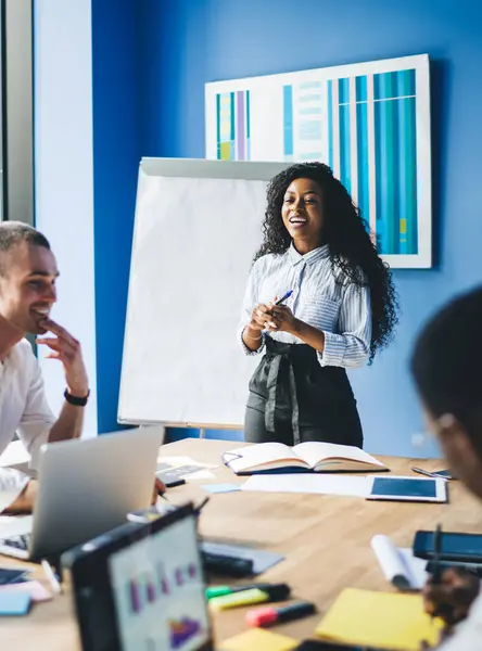 Content black woman talking to group of modern diverse coworkers at table meeting in conference room of new office with whiteboard
