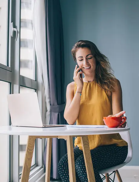 Happy young woman in yellow sleeveless blouse sitting at white round table with laptop and red mug communicating on smartphone at home
