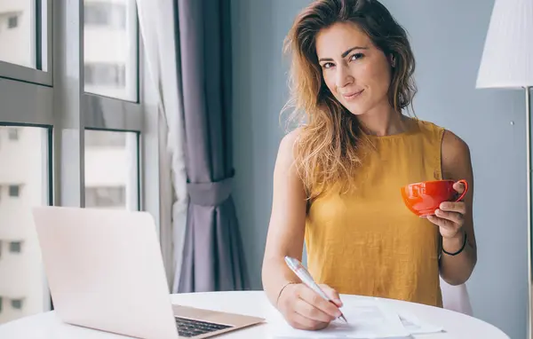 Coquettish young attractive woman in yellow sleeveless blouse sitting at white round table with laptop and red mug writing on paper at home and looking at camera