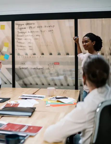 Executive black female in casual wear with marker discussing collaborative project with colleagues answering questions from coworker in modern office