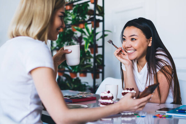 Multiethnic women in white t shirts texting on mobile phone sitting at table with cups of coffee and cakes in modern white studio