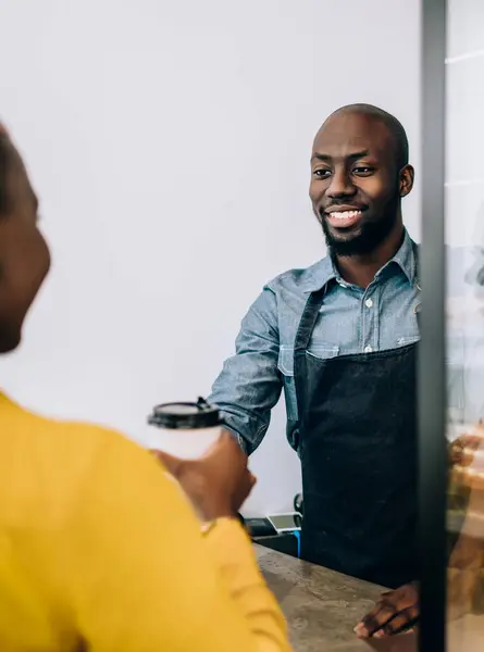 Smiling African American shop assistant in black apron standing at counter and giving disposable cup of coffee to crop customer in pastry shop