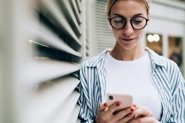 Gündelik kıyafetler giyen, şık gözlükler takan, akıllı telefon kullanan, duvara yaslanan ve son haberleri okuyan güzel bir bayan.