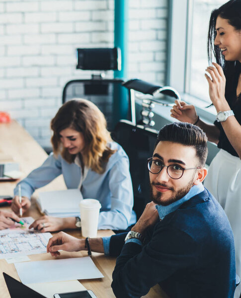 High angle of smiling diverse business partners in formal clothes examining blueprints and developing strategic plans at big wooden table with papers and devices