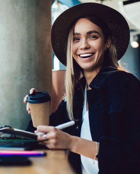 Excited young woman with clipboard and takeaway beverage smiling and looking at camera while sitting at cafe table and studying