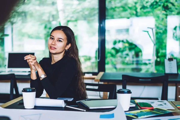 Young female designer in casual clothes sitting at table with papers and listening attentively to colleague while working together on design concept
