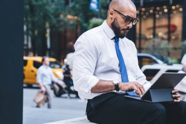 Kendine güveni tam, Afro-Amerikalı, şık giyinmiş, göz giysili, şehir merkezindeki bankta bulunan tabletin kablosuz klavyesine mesaj atan erkek.