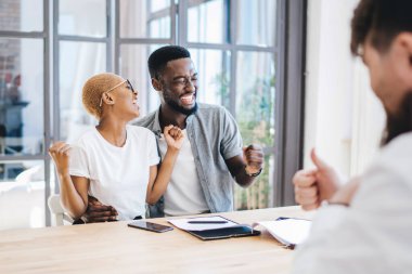 Sevgiyle kucaklaşan ve emlakçıyla aynı masada otururken yeni bir daire almayı kutlayan mutlu Afrikalı Amerikalı çift.