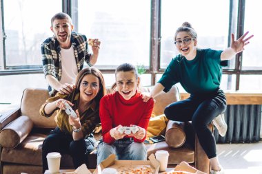 Oyun konsoluyla oynayan aktif, hareketli ve rahat bir grup insan oturma odasındaki kanepeye oturmuş pizza yiyor.
