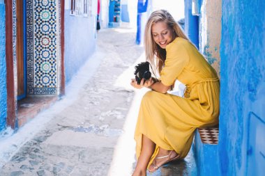 Fas 'ta güneşli bir günde mavi evin dışında oturmuş gülümseyen ve aşağı bakan iyimser kadın.