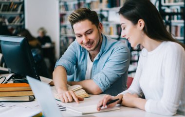 Çalışkan içerikli kadın ve erkek çalışırken gülüyor ve kütüphanede dizüstü bilgisayarlı açık kitaplarda bilgi arıyor.