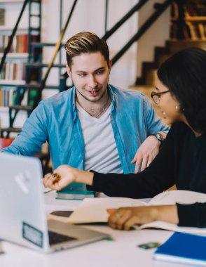Yakışıklı mutlu öğrenci, kütüphanede dizüstü bilgisayar kullanarak Afrikalı Amerikalı genç bir kadınla üniversite projesi yapıyor.