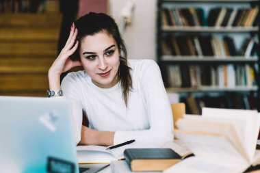 İçerik siyah saçlı beyaz ceketli bayan öğrenci masada oturuyor ve dizüstü bilgisayar ve kitaplarla ödev yaparken elini uzatıyor.