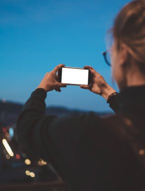 Tanımlanamayan sarışın fotoğrafçının elinde akıllı telefon tutarken ve boş ekrana bakarken gece şehrinin fotoğrafını çekerken.