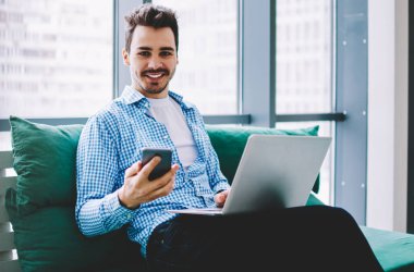 Neşeli gülümseyen adam dizüstü bilgisayarı bacaklarının üzerinde, partneriyle cep telefonu mesajlaşması kullanarak iş yerinde kameraya bakarak başarılı olmanın tadını çıkarıyor.