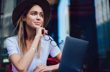 Şapkalı, gözlüklü, kafede otururken tablet kullanan, gülümseyen ve hayal kuran neşeli bir kadın.