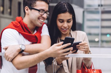 Moda tebessümlü, modern, çok ırklı genç adam ve kadın alışveriş çantasıyla akıllı telefonlara göz gezdirirken gün ışığında şehir kaldırımında duruyorlar.