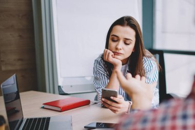 Kadın yöneticinin cep telefonuyla gündelik kıyafet sörfü yaparken masada oturması ve modern ofisteki erkek iş arkadaşıyla iletişim kurması yüksek açı.