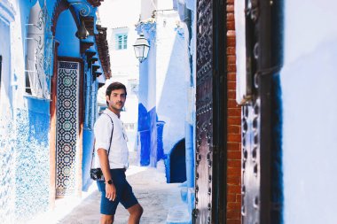 Fas 'ın ünlü şehri Chefchaouen' de mavi caddelerde dikilirken günlük kıyafetleriyle kameraya bakan düşünceli genç turistin yan görüntüsü.