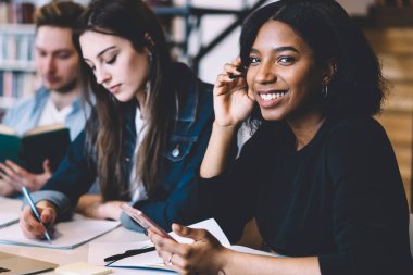 Mutlu, zeki, Afro-Amerikan kadın kameraya bakıyor ve rahat üniversite kütüphanesinde arkadaşlarıyla ders çalışırken cep telefonu kullanıyor.