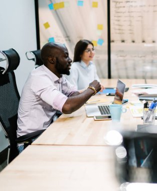 Beyaz gömlekli çok ırklı iş arkadaşları cam duvara karşı projeyi tartışırken dizüstü bilgisayar ve kağıtlarla masada oturuyorlar.