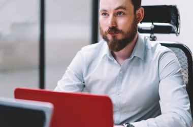 Ofis masaüstü önünde oturan meslektaşından sakallı ciddi erkek grafik tasarımcı dinleme bilgileri, çalışanları ile işbirliği sırasında dizüstü bilgisayar üzerinde çalışan genç adam. Yetişkin erkek işçi