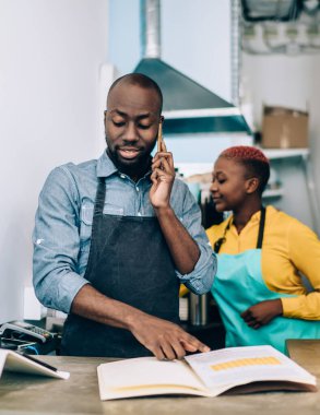 Önlüklü Afrikalı Amerikalı genç ve odaklanmış meslektaşlar cep telefonlarıyla konuşuyor. Masada ve tezgahta duruyorlar. Şehirdeki kafeteryada not defterlerini kontrol ediyorlar.