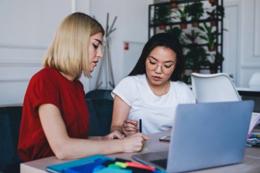 Genç ve zeki odaklı, gündelik giysiler içinde, dizüstü bilgisayarla çalışan, modern beyaz yaratıcı stüdyoda not defterine not alan, çok kültürlü kadınlar.