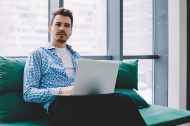 Düşünceli, genç, yakışıklı hippi gündelik giysiler içinde modern dizüstü bilgisayarla çalışıyor. İş yerinde bankta oturuyor ve başka tarafa bakıyor.