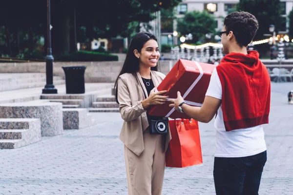 Side view of cheerful adult man in casual wear and glasses gives big gift box to excited smiling pretty young female with red shopping bag on square in city
