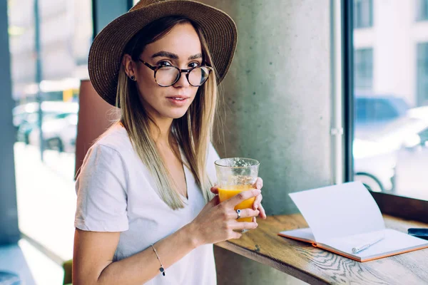 Adult modern female in casual outfit and with glasses wearing hat sitting at high table drinking juice while writing notes against glass window in cafe looking at camera