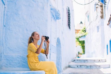 Çapraz bacaklı, neşeli genç sarışının yan görüntüsü. Fotoğraf makinesiyle caddedeki taş merdivenlerde oturan Fas 'ın eski binasını çekiyor.