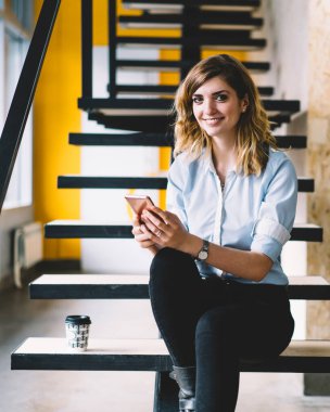 Ofis merdivenlerinde cep telefonu ile otururken kameraya bakan mutlu gülümseyen kadın çalışanın portresi, online alışveriş ve alışveriş için hücresel uygulama kullanarak resmi giyim pozitif kadın