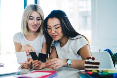 Modern neşeli kız arkadaşlar çalışma odasında masalarında oturup projeye hazırlanırken anlık fotoğraflara ve yapışkan notlara bakarlar.