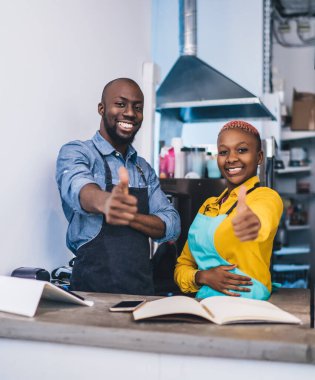 Gülümseyen Afro-Amerikan aşçılar sarı, gri gömlekli, mavi ve siyah önlüklü başparmak gösteren ve tezgahın arkasındaki kahkahalarla kameraya bakan