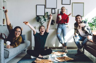 Heyecanlı genç grup, televizyonda maç izlerken ve rahat bir dairede pizza yerken farklı jestlerle başarıyı kutluyor.