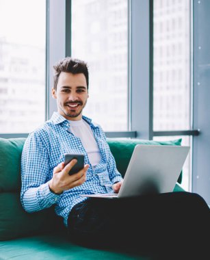 Neşeli gülümseyen adam dizüstü bilgisayarı bacaklarının üzerinde, partneriyle cep telefonu mesajlaşması kullanarak iş yerinde kameraya bakarak başarılı olmanın tadını çıkarıyor.