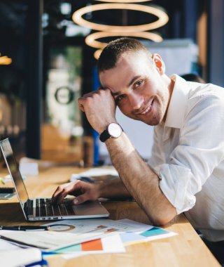 Çağdaş ofiste masa başında oturan içerikli adam, bulanık arkaplandaki kameraya bakarken dizüstü bilgisayar ve belgeler üzerinde çalışıyor.