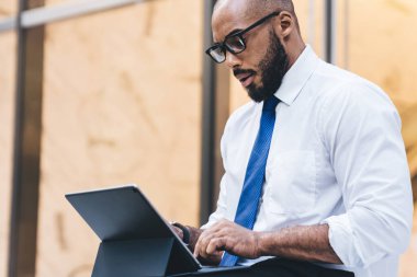 Afro-Amerikan iş adamının resmi giysileri içinde caddenin kenarında oturup tablet ekranına bakarken yan görünümü