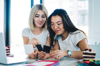 Modern neşeli kız arkadaşlar çalışma odasında masalarında oturup projeye hazırlanırken anlık fotoğraflara ve yapışkan notlara bakarlar.