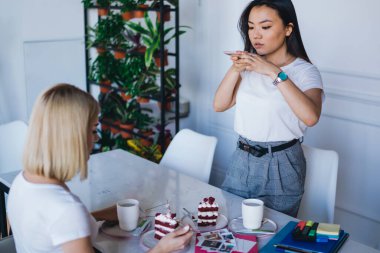Beyaz tişörtlü çok ırklı meslektaşlar, ellerinde kek fincanları, kahve fincanları, beyaz modern ofis alanındaki işaretler ve arka plandaki saksılarda bitkilerle masada oturuyorlar.