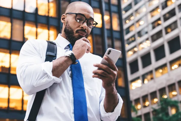 Gözlüklü, kel, düşünceli bir Afrikalı Amerikalı çalışanın modern ofis binalarının önünde dikilirken sakal okşaması ve beyaz akıllı telefonuyla mesajlaşması.