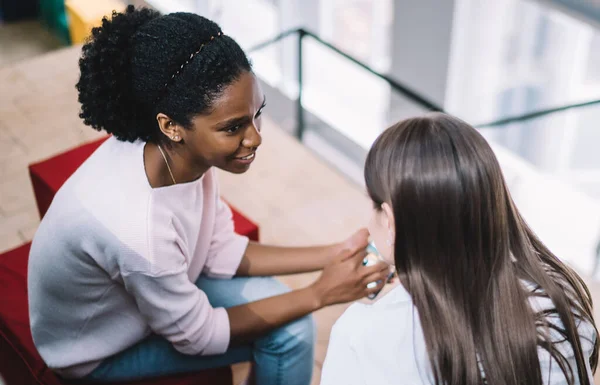 stock image From above satisfied young diverse women in casual clothes talking and sharing news while sitting together on poufs near stairs in college